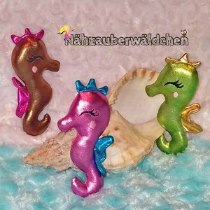 May include: Three plush seahorse ornaments with glitter accents. The seahorses are brown, pink, and green with gold crowns and blue fins. The seahorses are sitting on a seashell and a blue fuzzy surface.