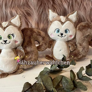 May include: Two plush brown and white squirrel toys with embroidered faces and blue eyes. The squirrels are sitting on a wooden surface with green leaves.