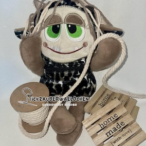May include: A brown and black plush toy with green eyes and a big smile. The toy is holding a spool of twine and is sitting on a wooden sign that says "home made [with love]"