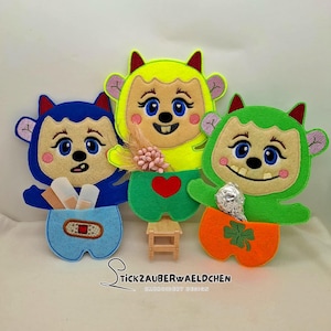 May include: Three felt monster-like figures in blue, yellow, and green. Each has a smiling face with blue eyes and a different item in their pockets: bandages, flowers, and a wrapped candy. The figures have horns and ears.