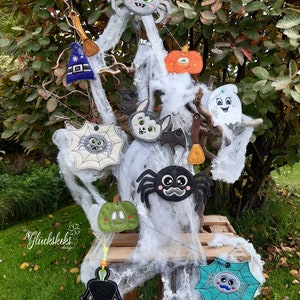 May include: A collection of Halloween-themed felt ornaments, including a spider, a ghost, a pumpkin, a bat, a witch's hat, and a broom. The ornaments are hanging from a white spider web on a wooden crate.