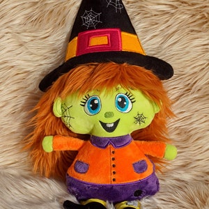 May include: A green and orange plush witch doll with orange hair, a black witch hat with a spiderweb design, and a purple skirt. The doll is sitting on a fuzzy brown surface.