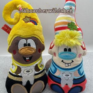 May include: Two plush gnomes with embroidered faces and teeth. The gnome on the left is wearing a yellow hat with a rainbow and a star. The gnome on the right is wearing a red and white striped hat with a shamrock. Both gnomes are wearing black pants and have a white tooth in their hands. The text "Nähzauberwäldchen" is embroidered on the top of the image.