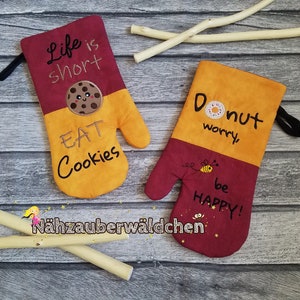 May include: Two oven mitts with embroidered sayings. The first mitt is yellow and red with the text "Life is short Eat Cookies" and a smiling cookie. The second mitt is yellow and red with the text "Donut worry be happy!" and a bee.
