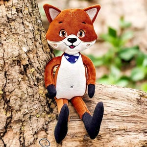May include: A plush fox doll with brown fur, white accents, and a blue bandana. The fox has black paws and is sitting on a tree branch. The doll has the text "Stickzauberwaelchen Embroidery Design" at the bottom.