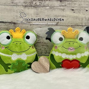 Puede incluir: Dos muñecos de peluche verdes de rana príncipe y princesa con ojos grandes, con coronas doradas. El príncipe tiene una pajarita blanca y un trébol, mientras que la princesa sostiene un corazón rojo. Un corazón de madera está entre ellos. El texto "Stickzauberwaeldchen Embroidery Design" está arriba.
