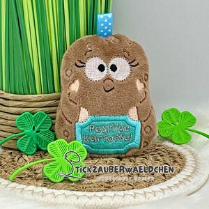 May include: A brown plush potato-shaped toy with embroidered facial features and the words "Positive Kartoffel" on a teal plaque. Green shamrock decorations and a blue polka-dot ribbon are also visible.