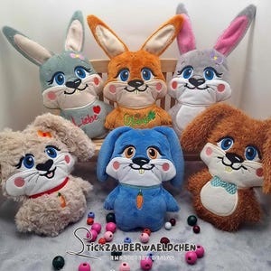 Puede incluir: Cinco juguetes de peluche de conejo con detalles bordados. Los conejos son de varios colores, incluyendo gris, naranja, azul y marrón. Cada conejo tiene un diseño bordado diferente en su pecho. Los conejos están sentados sobre una superficie blanca.