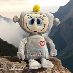 Könnte beinhalten: Ein graues Plüschroboter-Spielzeug mit einem Herzen auf der Brust und einem blau-grauen EKG-Muster auf dem Bauch. Der Roboter hat eine gelbe Antenne und große blaue Augen. Der Roboter sitzt auf einem Felsen mit einem Berg im Hintergrund.