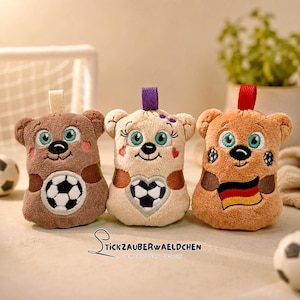 Op de afbeelding: Drie pluche berenspeelgoed met voetbalontwerpen. De beren zijn bruin, beige en oranje, elk met een lus in een andere kleur aan de bovenkant. De beren hebben voetbalontwerpen op hun buik en wangen. De beren hebben groene ogen en lachen.