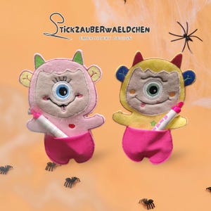 May include: Two felt monster-shaped pen holders in pink and yellow, each with a single eye and holding a pen. The text "Stickzauberwaeldchen Embroidery Design" is at the top. Spiders and spiderwebs are in the background.