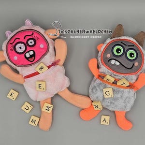 May include: Two plush toys with zippered pockets, each holding letter tiles from a Scrabble game. The pink toy has a playful expression with large eyes and a wide mouth. The gray toy has a grumpy expression with green eyes and a frown. The text "TICK ZAUBERWAELDCHEN EMBROIDERY DESIGN" is visible in the background.