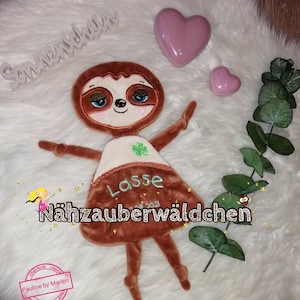 Puede incluir: Un juguete de peluche de perezoso marrón con un trébol verde en el pecho. El perezoso lleva una camisa blanca con el nombre "Lasse" bordado. El juguete está sentado sobre una superficie blanca y esponjosa con un corazón rosa y una rama de eucalipto.