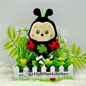 Könnte beinhalten: Eine rot-schwarze Marienkäfer mit einem grünen Kleeblatt auf der Brust, gestickt auf einem weißen Lattenzaun mit grünem Gras und Blumen. Der Text "Stickzauberwaeldchen Embroidery Design" befindet sich unter dem Zaun.