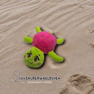 Peut inclure: Une peluche en forme de tortue avec une carapace rose, des membres verts et une tête verte souriante. Le jouet est posé sur une surface sablonneuse. Le texte "StickZauberwaeldchen Embroidery Design" est visible.