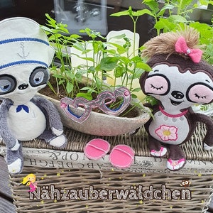 May include: Two plush sloth toys, one wearing a sailor hat and the other with a pink bow, are sitting on a wicker basket with a plant in the background. The basket has a newspaper print on the bottom and the text "Nähzauberwäldchen" on the front.