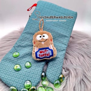 May include: A brown plush toy with a smiling face and the text "Mut-Toffel" on a blue banner, hanging from a teal oven mitt. The mitt has a geometric pattern and a red loop. Green glass beads are scattered around the mitt.