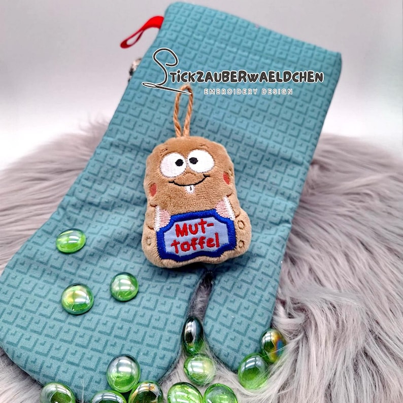 May include: A brown plush toy with a smiling face and the text "Mut-Toffel" on a blue banner, hanging from a teal oven mitt. The mitt has a geometric pattern and a red loop. Green glass beads are scattered around the mitt.