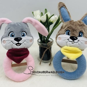 Puede incluir: Dos juguetes de sonajero de peluche en forma de conejo. Uno es gris con orejas rosas y una bufanda roja, el otro es marr&oacute;n con orejas azules y una bufanda amarilla. Ambos tienen caras bordadas y un anillo de madera en el centro.