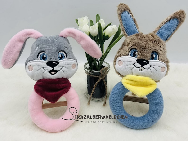 Puede incluir: Dos juguetes de sonajero de peluche en forma de conejo. Uno es gris con orejas rosas y una bufanda roja, el otro es marr&oacute;n con orejas azules y una bufanda amarilla. Ambos tienen caras bordadas y un anillo de madera en el centro.