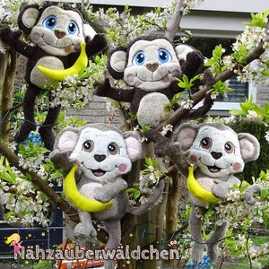 May include: Four plush monkey toys with big blue eyes and toothy grins are holding yellow bananas. The monkeys are sitting on a tree branch with white blossoms. The text "Nahzauberwäldchen" is visible at the bottom of the image.