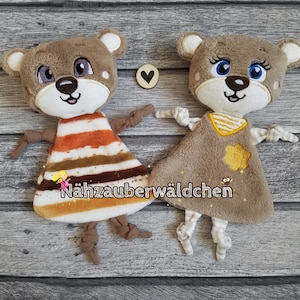 May include: Two brown plush teddy bear toys with embroidered faces and knotted limbs. One bear is wearing a brown and orange striped dress and the other is wearing a brown dress with a yellow flower and stripes. The text "Nähzauberwäldchen" is embroidered on the brown dress.
