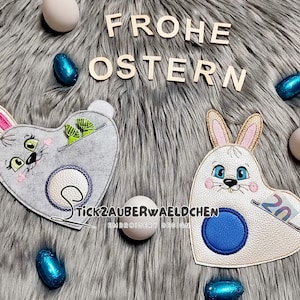 May include: Two heart-shaped Easter bunny-themed felt pouches. One is gray with green accents, the other is white with blue accents. The words "FROHE OSTERN" are displayed above the pouches. The pouches have a coin slot and a pocket.