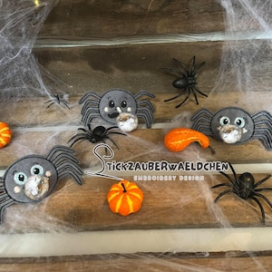 Puede incluir: Un conjunto de cinco aplicaciones de araña de fieltro gris con detalles bordados. Las arañas tienen ojos saltones y están rodeadas de calabazas naranja y amarillas y una telaraña.