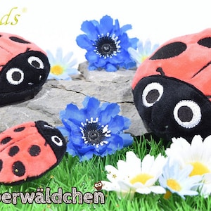 Puede incluir: Tres juguetes de peluche de mariquita de diferentes tamaños, con manchas negras y ojos blancos, están dispuestos en una roca con hierba verde y flores blancas y azules.