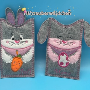 May include: Two gray felt bunny-shaped pouches with pink and white embroidery. One bunny holds a carrot with a smiling face and the name "Juli" embroidered below. The other bunny holds a pink and white Easter egg and the name "Mama" embroidered below.