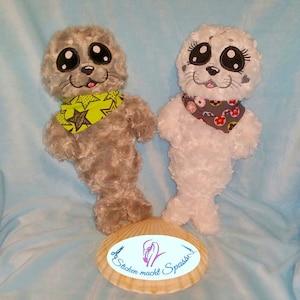 May include: Two plush toy seals with big eyes and a smiling expression. The seal on the left is gray with a green and black star patterned scarf. The seal on the right is white with a gray and red polka dot scarf. The seals are sitting on a seashell with the text "Sticken macht Spass".
