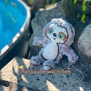 Puede incluir: Un juguete de peluche de perezoso marrón y gris con grandes ojos verdes y una expresión feliz. El perezoso está sentado en una roca cerca de una piscina.