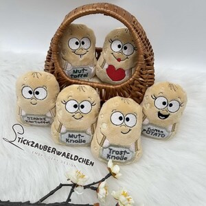 May include: A selection of plush potato-shaped toys with embroidered faces and small signs. Some are in a woven basket. The signs have German text. The toys are beige with black and white details.