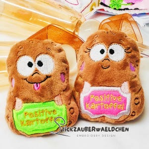 May include: Two plush potato-shaped toys with embroidered faces and holding signs. One sign is green and reads "Positive Kartoffel", the other is pink and reads the same. The toys are brown with pink accents and black and white eyes.