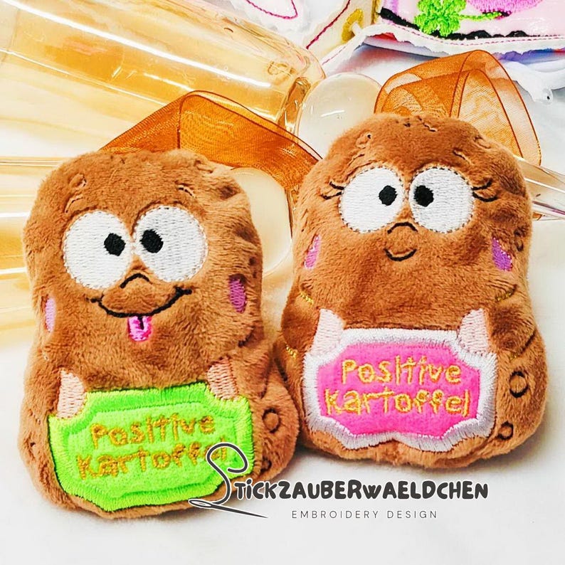 May include: Two plush potato-shaped toys with embroidered faces and holding signs. One sign is green and reads "Positive Kartoffel", the other is pink and reads the same. The toys are brown with pink accents and black and white eyes.