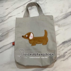 May include: A gray canvas tote bag with a brown dachshund embroidered on the front. The dachshund has pink ears with white flowers. The bag has the text "Stickzauberwaeldchen Embroidery Design" on the bottom.