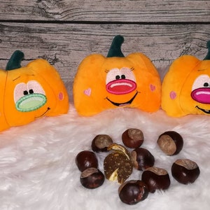 Könnte beinhalten: Drei orangefarbene Plüschkürbisse mit gestickten Gesichtern, zwei mit rosa Nasen und einer mit grüner Nase, sind auf einer weißen, flauschigen Oberfläche mit braunen Kastanien um sie herum angeordnet. Die Kürbisse haben gestickte Details und einen grünen Stiel. Der Hintergrund ist eine Holzoberfläche.