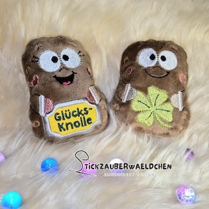 May include: Two plush, brown potato-shaped toys with embroidered faces and details. One has a yellow tag with text, the other a green four-leaf clover. The toys are on a fluffy, white surface.