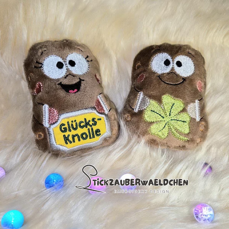 May include: Two plush, brown potato-shaped toys with embroidered faces and details. One has a yellow tag with text, the other a green four-leaf clover. The toys are on a fluffy, white surface.