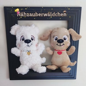 May include: Two plush dog toys, one white and one brown, are displayed in a black frame. The white dog has a black nose and brown eyes, while the brown dog has a black nose and blue eyes. The frame has the text "Nähzauberwäldchen" in gold lettering.