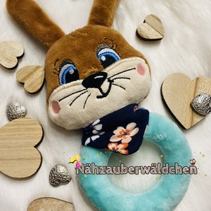 Puede incluir: Un sonajero de peluche marr&oacute;n en forma de conejo con ojos azules y un pa&ntilde;uelo con estampado floral. El sonajero es turquesa y tiene forma de anillo. El texto "N&auml;hzauberw&auml;ldchen" es visible en el anillo.