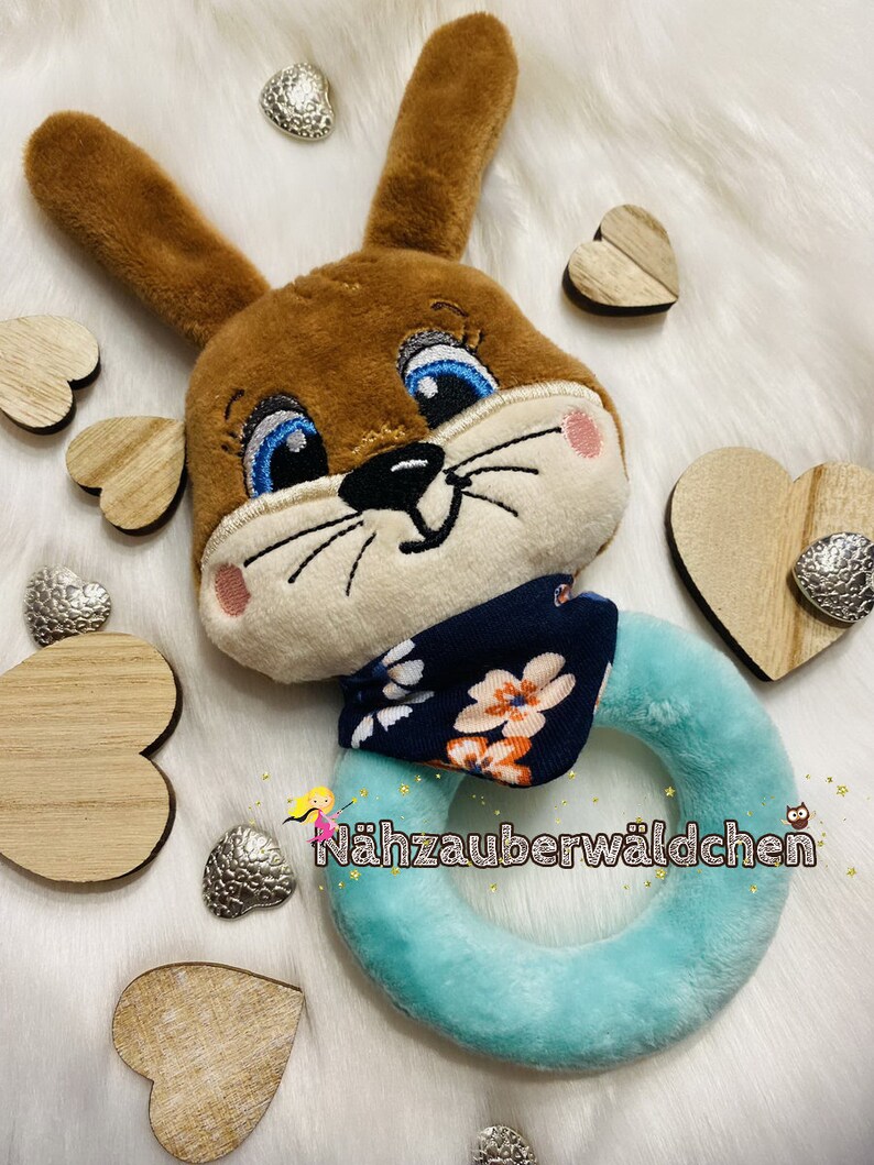 Puede incluir: Un sonajero de peluche marr&oacute;n en forma de conejo con ojos azules y un pa&ntilde;uelo con estampado floral. El sonajero es turquesa y tiene forma de anillo. El texto "N&auml;hzauberw&auml;ldchen" es visible en el anillo.