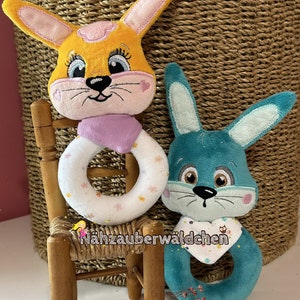 Puede incluir: Dos juguetes de peluche con forma de conejo, uno amarillo con orejas rosas y un anillo blanco y rosa, el otro azul turquesa con orejas azul claro y un anillo blanco y turquesa. Los juguetes est&aacute;n sentados en una peque&ntilde;a silla de madera.