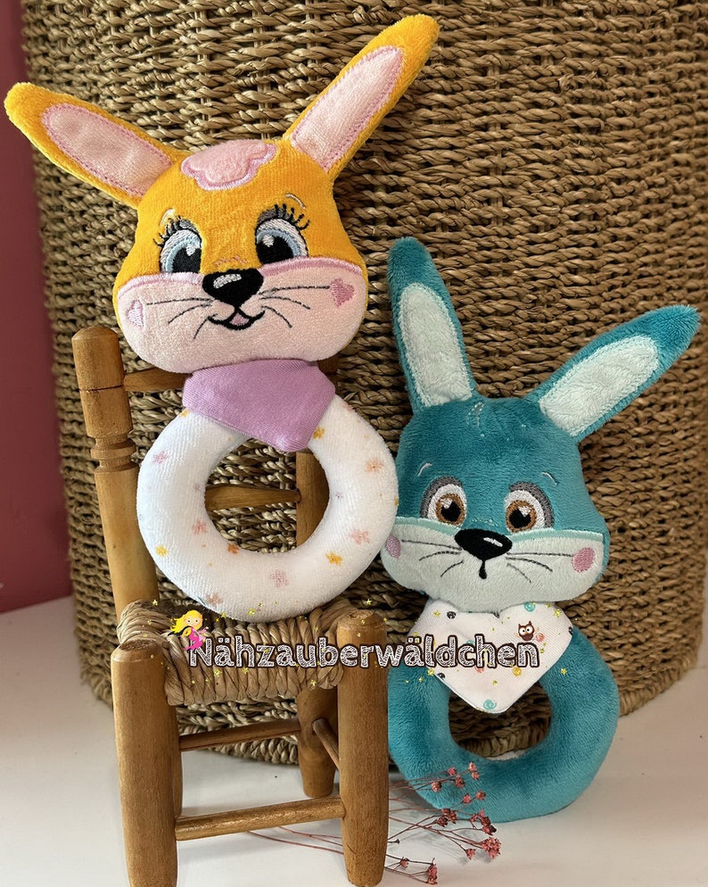 Puede incluir: Dos juguetes de peluche con forma de conejo, uno amarillo con orejas rosas y un anillo blanco y rosa, el otro azul turquesa con orejas azul claro y un anillo blanco y turquesa. Los juguetes est&aacute;n sentados en una peque&ntilde;a silla de madera.