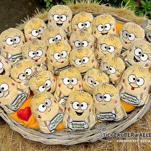 May include: A wicker basket overflowing with plush potato toys. Each toy features embroidered eyes, eyelashes, and a smiling mouth. Some have a small sign that reads "Positive Potato" or "Gl&uuml;cks Knolle". The basket rests on straw.
