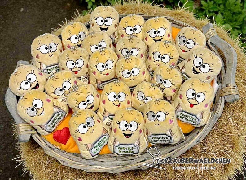 May include: A wicker basket overflowing with plush potato toys. Each toy features embroidered eyes, eyelashes, and a smiling mouth. Some have a small sign that reads "Positive Potato" or "Gl&uuml;cks Knolle". The basket rests on straw.