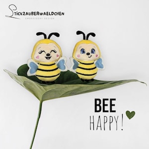 Puede incluir: Dos juguetes de peluche de abeja con cuerpos a rayas amarillas y negras, alas azules y caras sonrientes, descansando sobre una gran hoja verde. La frase "BEE HAPPY!" está escrita debajo con un corazón verde.