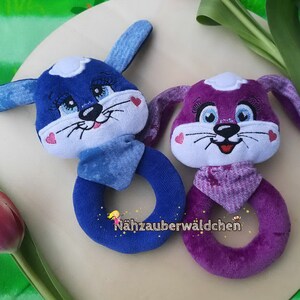 Puede incluir: Dos sonajeros de peluche con forma de animal, uno azul y otro morado, con caras bordadas y un anillo para que el beb&eacute; los sostenga. El sonajero azul tiene una bufanda blanca y azul y el sonajero morado tiene una bufanda blanca y morada. El texto "N&auml;hzauberw&auml;ldchen" es visible en la parte inferior de la imagen.