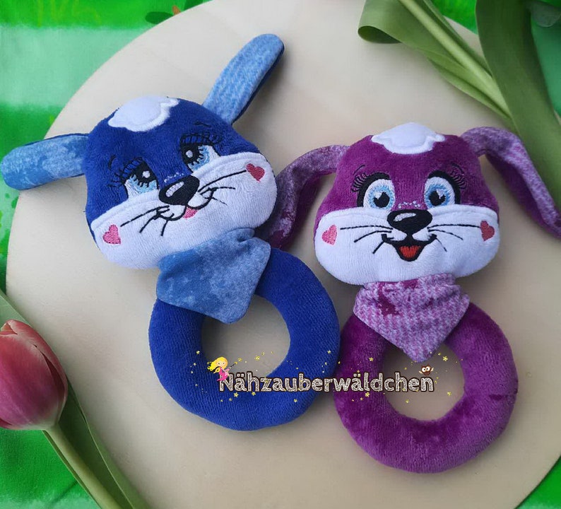 Puede incluir: Dos sonajeros de peluche con forma de animal, uno azul y otro morado, con caras bordadas y un anillo para que el beb&eacute; los sostenga. El sonajero azul tiene una bufanda blanca y azul y el sonajero morado tiene una bufanda blanca y morada. El texto "N&auml;hzauberw&auml;ldchen" es visible en la parte inferior de la imagen.