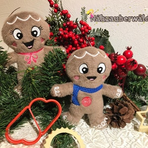 May include: Two plush gingerbread people ornaments with smiling faces, brown fur, and embroidered details. The ornaments are surrounded by green pine branches, red berries, and a pine cone.